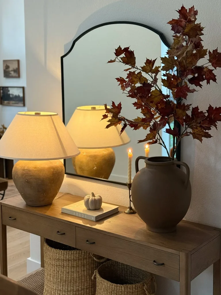 A wooden console table styled for Thanksgiving with a lamp, a vase of autumn leaves, two candlesticks, a small decorative pumpkin, and a mirror above, while baskets are neatly stored underneath.