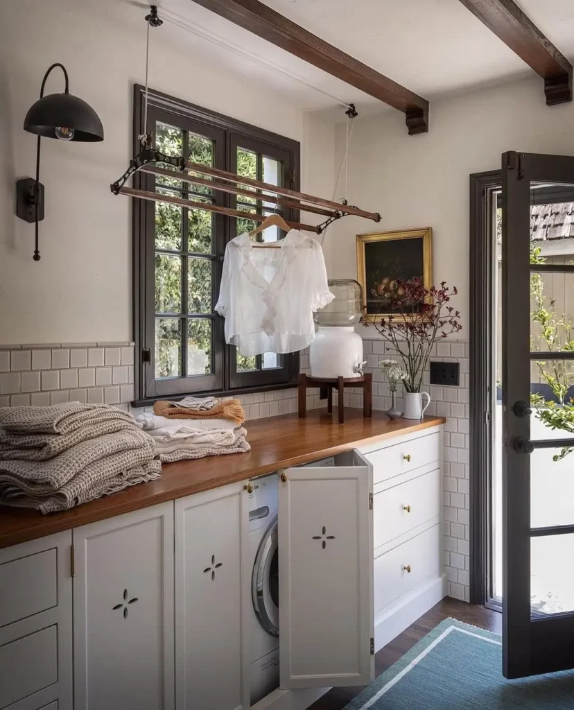Charming laundry room with a wooden countertop, built-in washer, wall-mounted drying rack, folded linens, vintage wall art, and natural light from large windows.