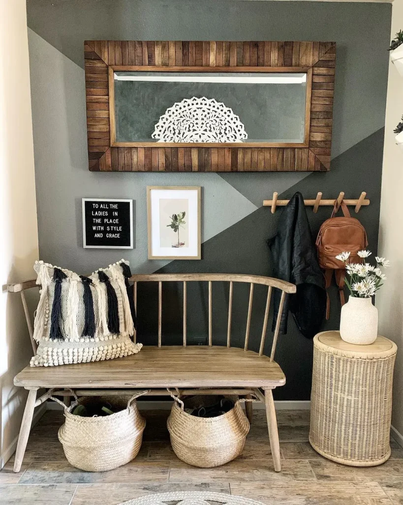 A wooden bench with pillows and baskets sits against a wall with a mirror, framed art, a quote sign, coat hooks, jackets, and a round wicker side table topped with a vase of flowers.
