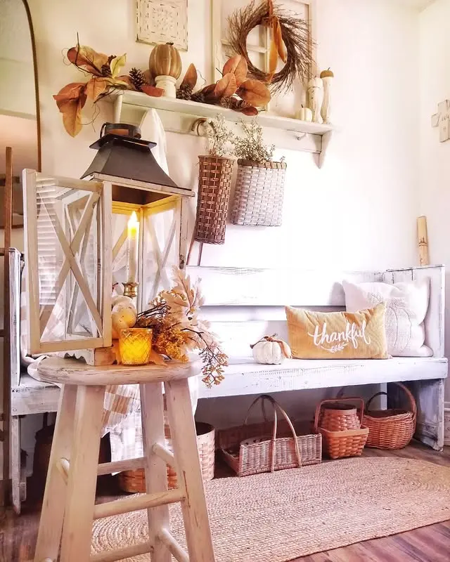 A cozy, rustic entryway decorated with autumn-themed items, woven baskets, a "thankful" pillow on a white bench, and warm fall lighting from a lantern with a lit candle on a stool.
