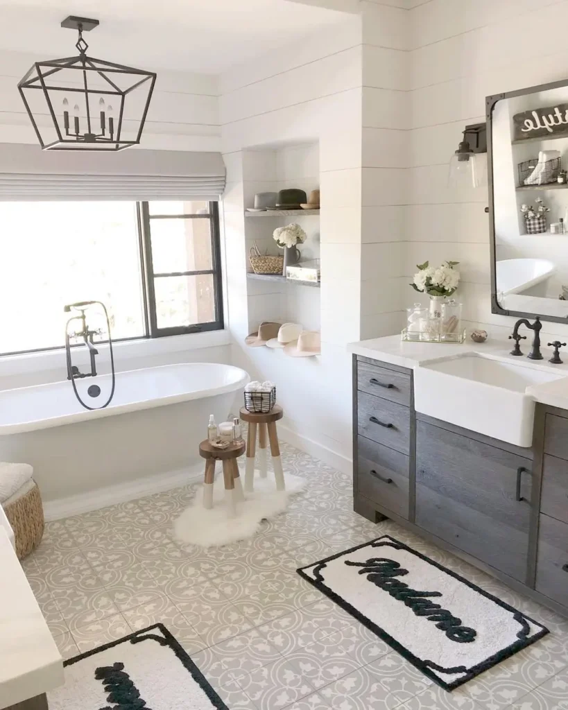Modern farmhouse bathroom with a freestanding tub, gray vanity, large mirror, shelves with decor, patterned floor tiles, and two rugs labeled "Madame" and "Monsieur.