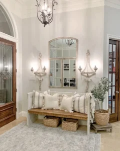 A light-filled farmhouse entryway with a wooden bench, striped pillows, woven baskets underneath, a mirror above, wall sconces, a potted plant, and a chandelier overhead.