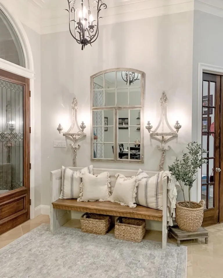 A light-filled farmhouse entryway with a wooden bench, striped pillows, woven baskets underneath, a mirror above, wall sconces, a potted plant, and a chandelier overhead.