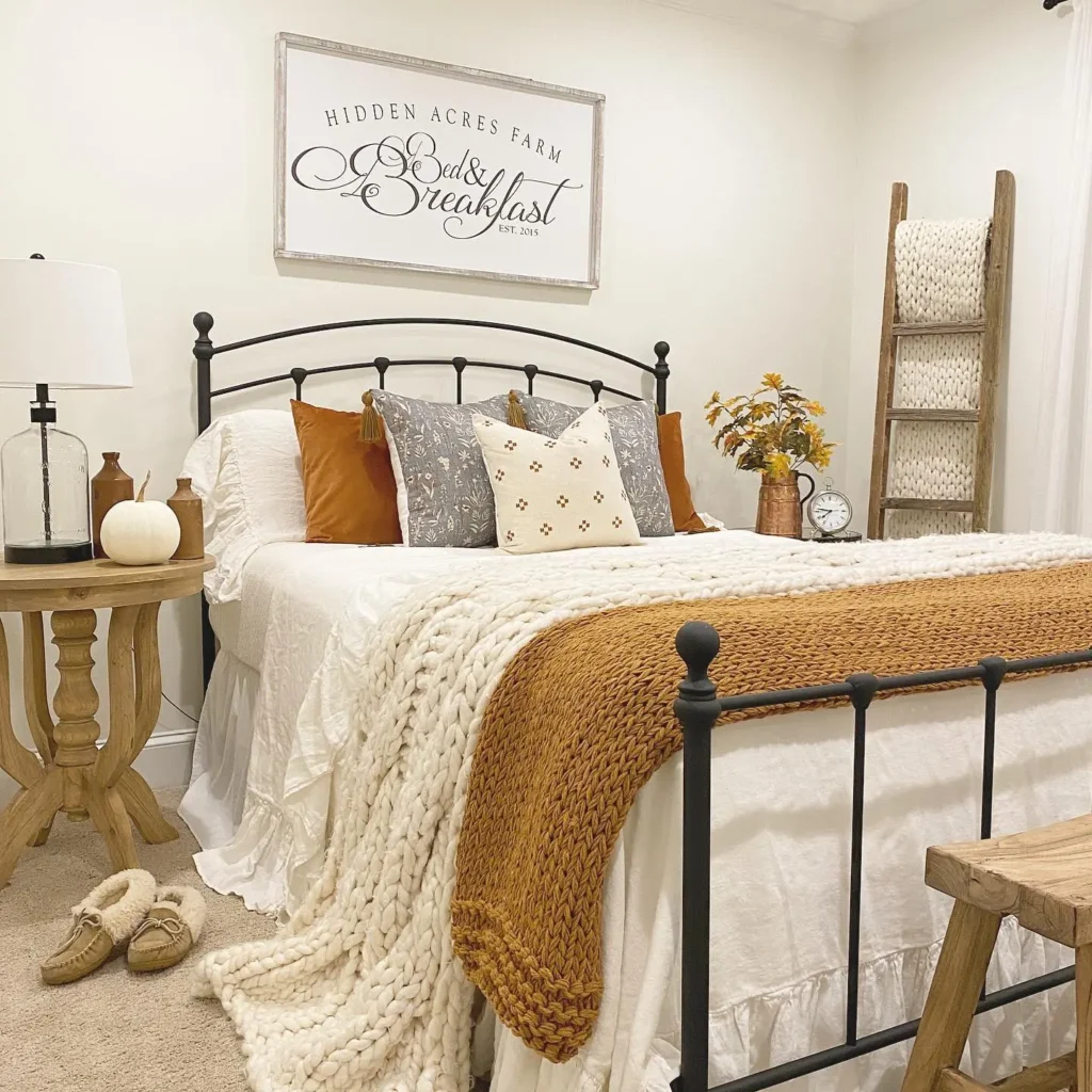 A cozy farmhouse bedroom features a black metal bed frame, white bedding with textured blankets, brown accent pillows, and subtle fall touches on the nightstand, a decorative sign on the wall, and a bedside table with a lamp.