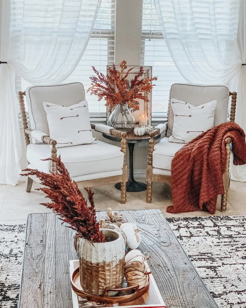 Two cushioned wooden chairs with white pillows face a small round table holding a vase of autumn leaves, a candle with a cozy fall scent, and small pumpkins. A red throw blanket is draped over one chair.