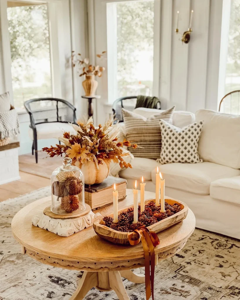 A cozy farmhouse fall living room with a round wooden coffee table decorated with lit candles, pinecones, and autumnal floral arrangements, surrounded by neutral-toned furniture and pillows.