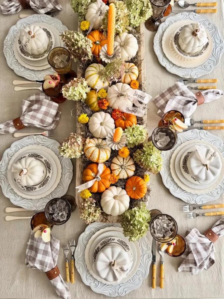Table set for a fall-themed meal with plates, cutlery, brown glasses, plaid napkins, and a farmhouse-inspired centerpiece of small pumpkins, gourds, flowers, and a tall candle styled in a wooden crate.