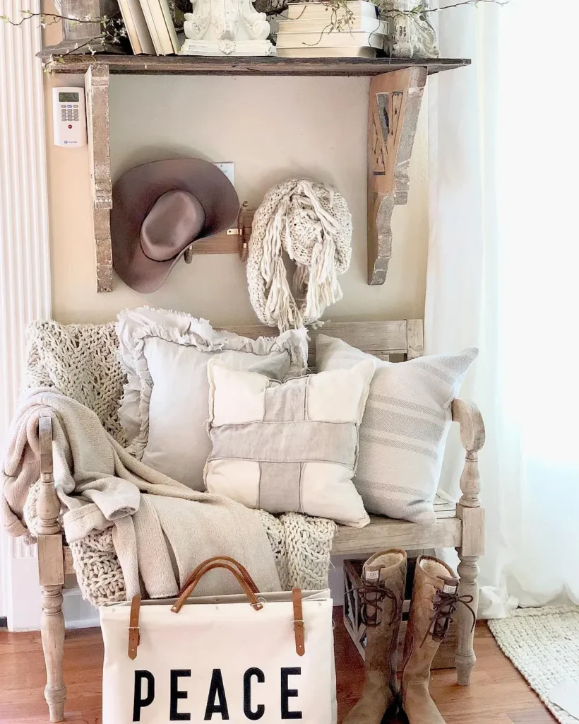A rustic wooden bench with farmhouse neutral pillows and blankets sits under a wall shelf holding decor items. Boots, a hat, a knit scarf, and a bag labeled "PEACE" are nearby.