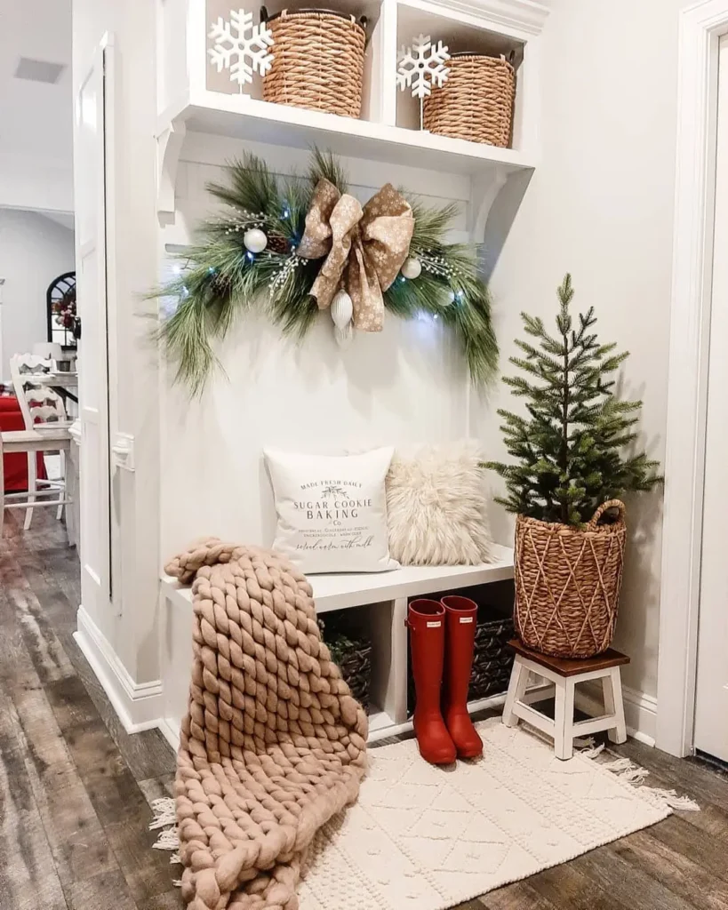 A hallway bench decorated for the holidays with farmhouse mixed pillows, a garland, bow, and pine branches, red rain boots, a chunky knit blanket, a small fir tree, and woven baskets on shelves.