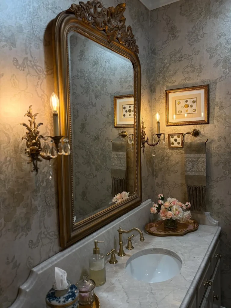 Elegant bathroom featuring a French country mirror with ornate gold frame, marble sink, gold fixtures, floral arrangement, and decorative wall sconces providing warm lighting.