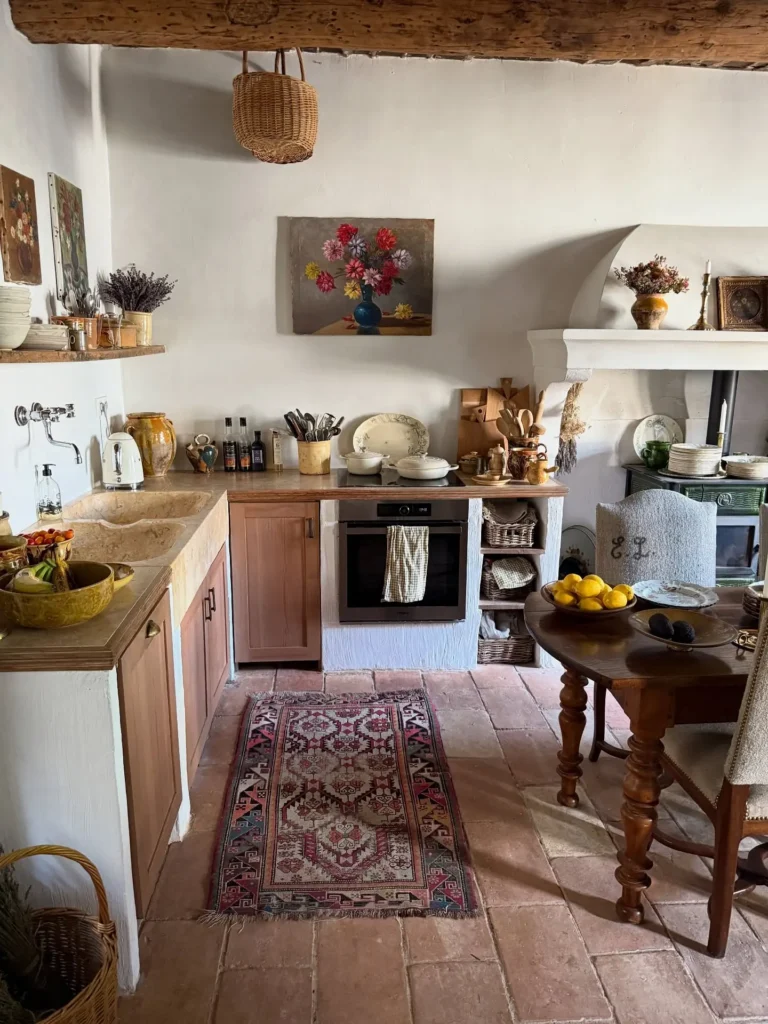 A rustic French country kitchen with wooden cabinets, a small oven, open shelves, a rug, a table with lemons, and wall art, featuring earthy tones and vintage decor.