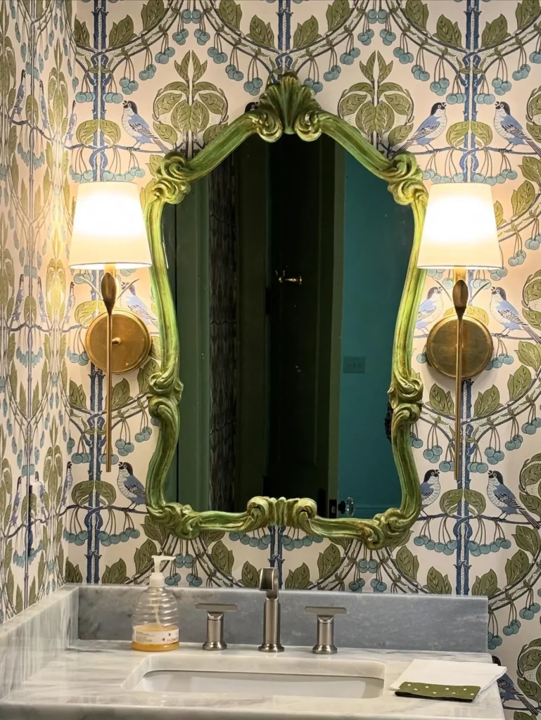 A vintage-style french country bathroom with a green ornate mirror, two wall sconces, floral wallpaper, a marble sink, and a soap dispenser on the counter.
