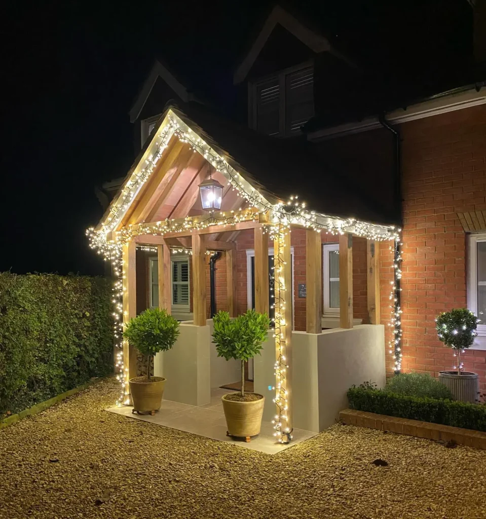 A front porch decorated with string lights at night, featuring two potted plants at the entrance and gravel surrounding the area.