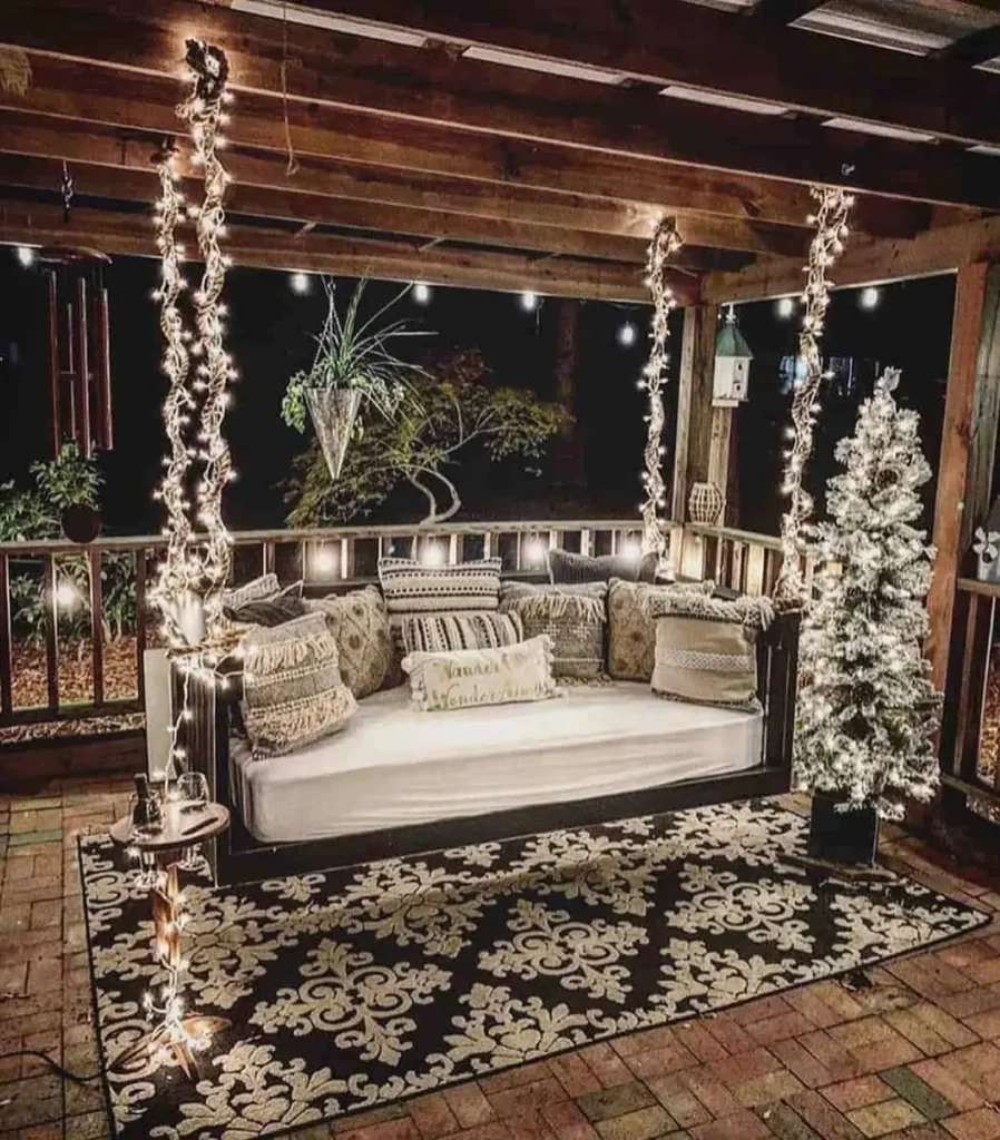 A covered outdoor patio with a cushioned swing sofa, decorative pillows, glowing string lights overhead, a white artificial Christmas tree, and a patterned rug.