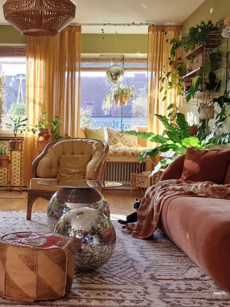 A cozy, funky bohemian living room with warm-toned furniture, large plants, two disco balls on a rug, sunlight streaming through yellow curtains, and a black-and-white cat lounging near the sofa.