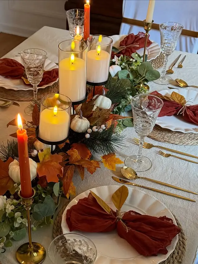 Elegant fall table setting featuring a centerpiece garland with greenery, orange leaves, and mini white pumpkins. The table is styled with pillar candles in glass holders, gold flatware, crystal glasses, and rust-colored napkins tied with gold leaves on white plates for a warm autumn ambiance.