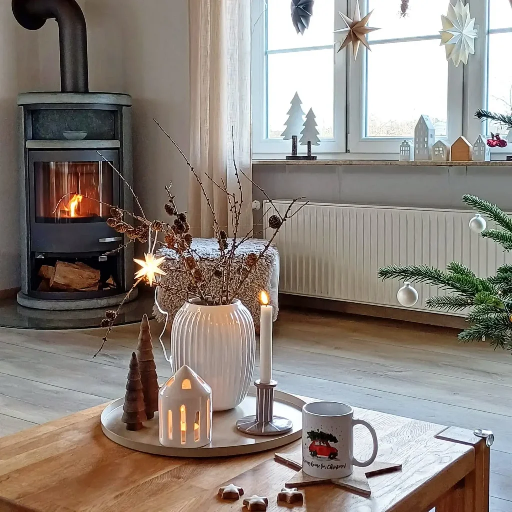 A cozy, hygge winter living room with a lit wood stove, a decorated coffee table featuring a vase, candle, mug, and wooden ornaments, nestled beside a Christmas tree and festive window decorations.