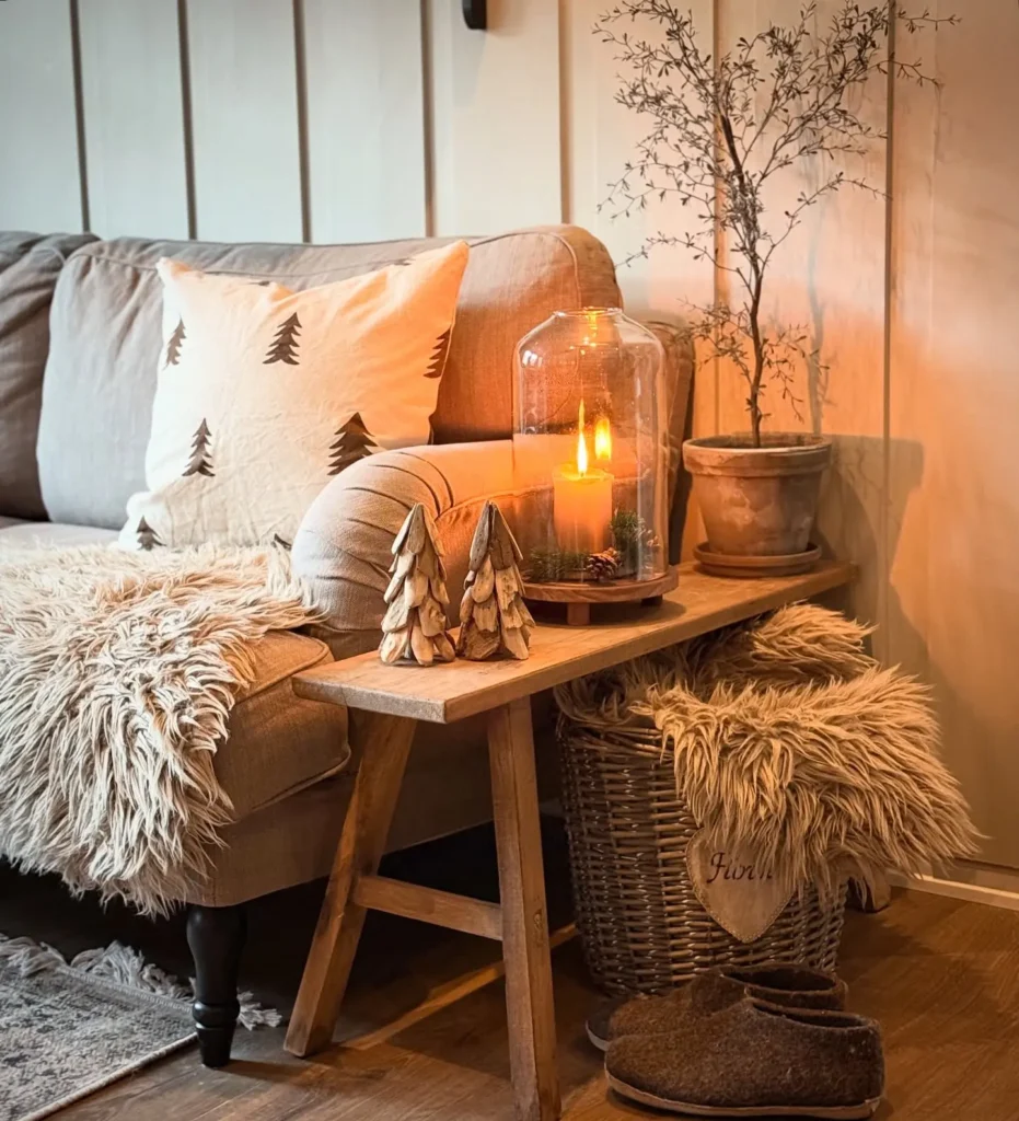 A cozy hygge winter living room corner with a beige sofa, tree-patterned pillow, fur throw, wooden bench with candles, small tree ornaments, potted plant, basket, and slippers on a wooden floor.