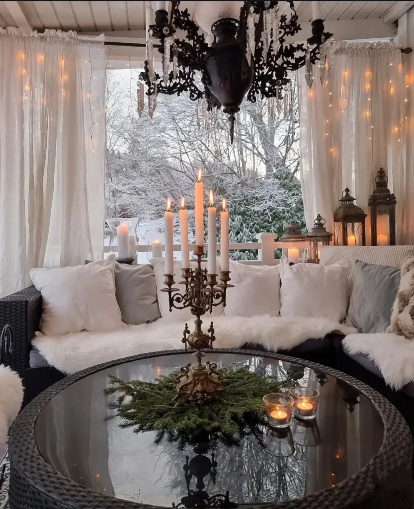 A cozy hygge winter outdoor seating area with white cushions, a glass table adorned with candles and greenery, string lights twinkling above, and snowy trees visible through sheer curtains.