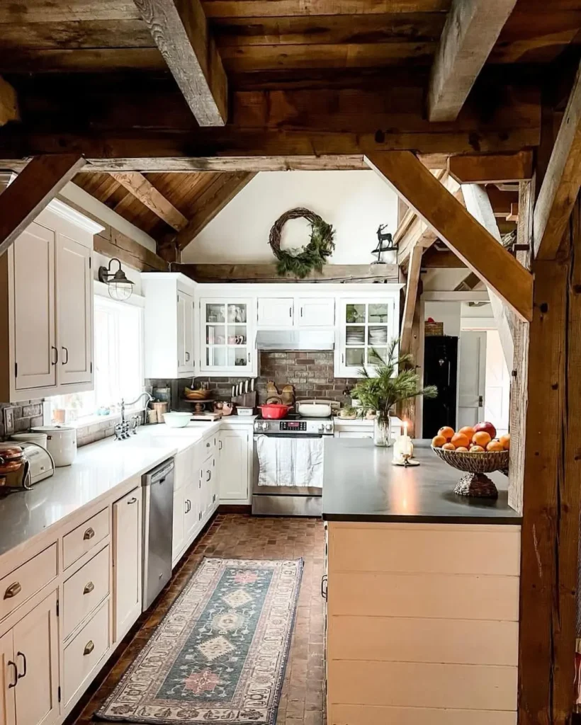 A charming rustic kitchen with white cabinets, exposed wooden beams, a patterned rug, stainless steel appliances, and a wreath hanging above the stove.