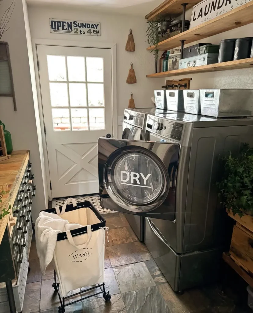 A modern laundry room designed to maximize laundry room storage features washer and dryer, open shelves, metal storage bins, a rolling cart, and a door with a window.