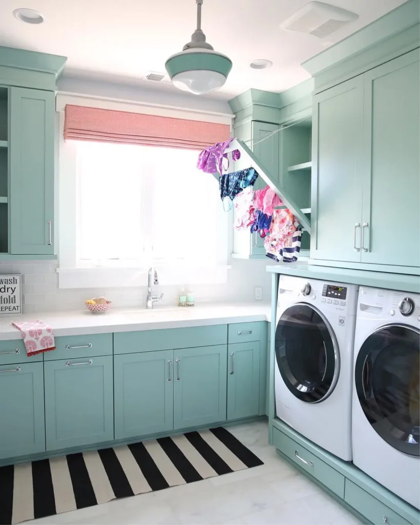 Bright laundry room with aqua cabinetry, front-loading washer and dryer, built-in drying rack, white countertops, striped rug, and a pink Roman shade over the sink.
