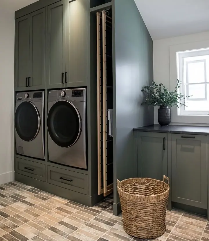 Modern laundry room with green built-in cabinets, stacked front-loading washer and dryer, pull-out vertical storage, dark countertop, woven laundry basket, and natural light from a window.