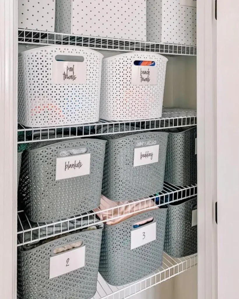 Wire shelves in a closet hold labeled white and gray storage bins for laundry, pool towels, beach towels, blankets, bedding, and numbered bins for organized storage.