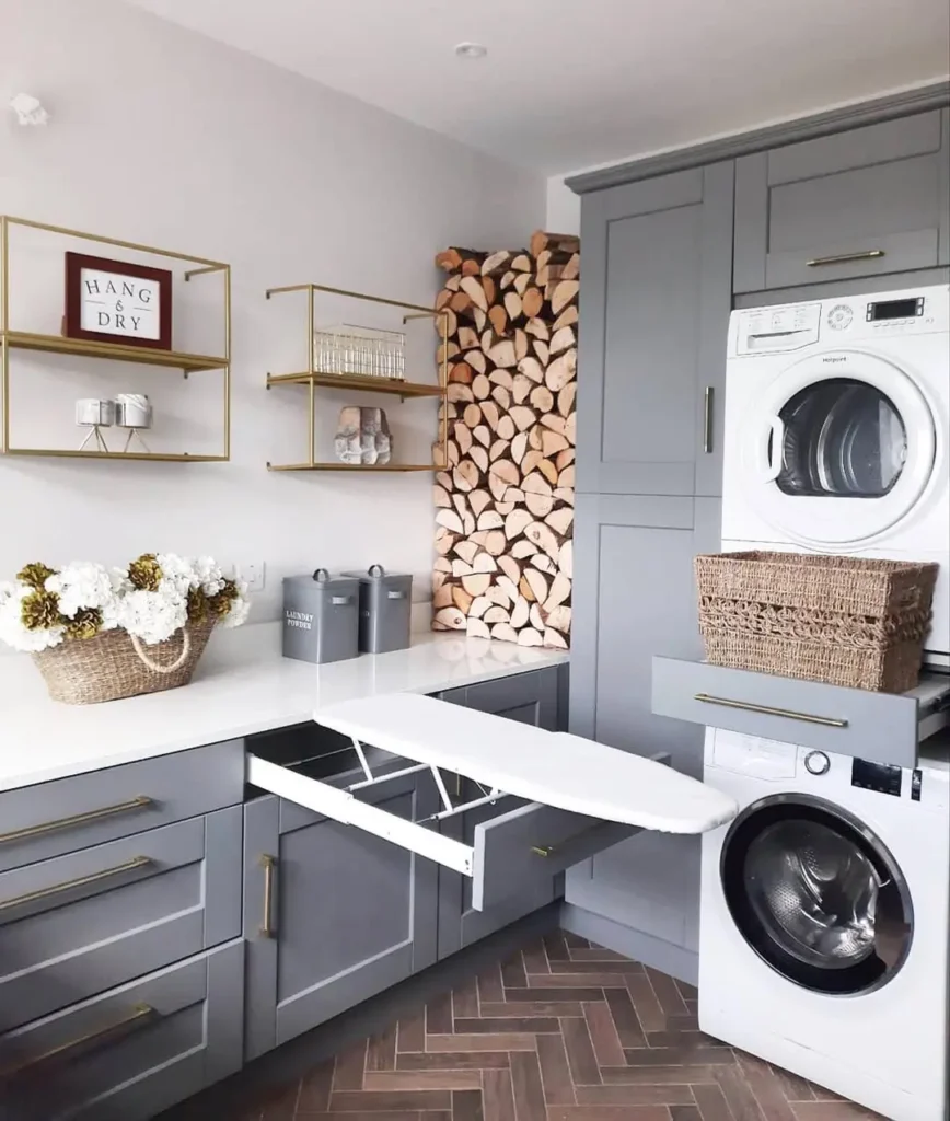 A modern laundry room with stacked washer and dryer, creative solutions like a pull-out ironing board, shelves, a basket of flowers, and tidy firewood stacked in the corner.