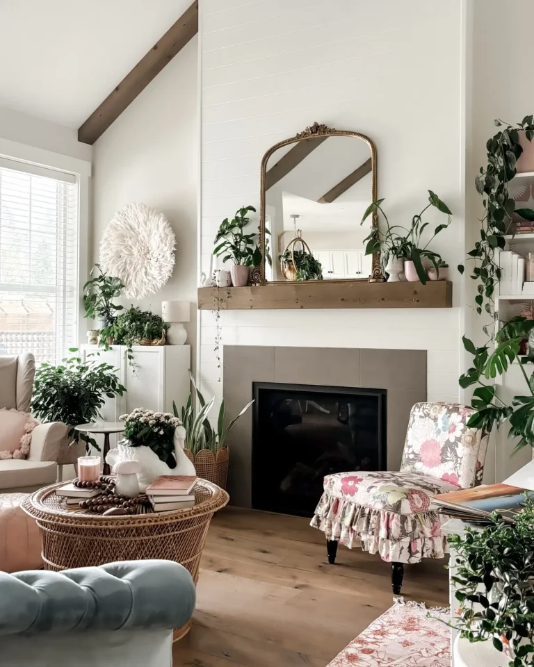 Bright and cozy living room decorated with indoor plants, a wooden mantel with a vintage gold mirror, wicker coffee table styling, and a floral accent chair creating a warm, nature-inspired home decor look.