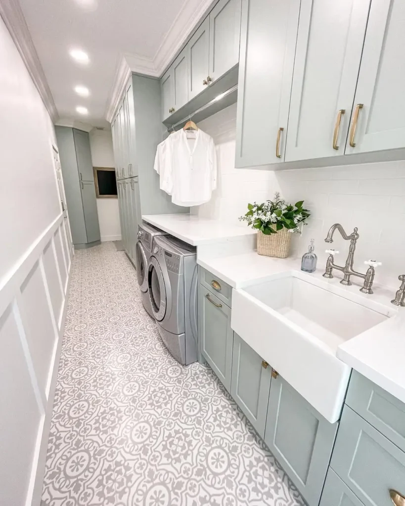 A modern, long narrow laundry room with light blue cabinets, patterned tile floor, washer and dryer, farmhouse sink, and a white shirt hanging above the counter.