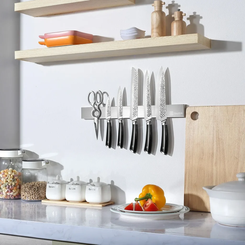 A modern kitchen counter with a magnetic knife strip holding several knives and scissors, shelves lined with jars, a cutting board displaying sliced bell pepper, and organized spice containers.
