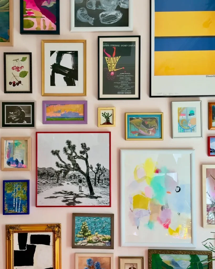 A variety of framed artworks, including maximalist abstract paintings, landscapes, and photographs, are arranged in a gallery style on a light-colored wall.