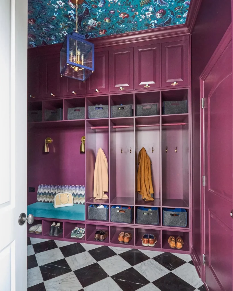 A vibrant maximalist mudroom featuring deep plum built-in lockers, a bold teal patterned ceiling, brass wall sconces, an ikat bench cushion, checkerboard marble flooring, and mixed textures that create a dramatic and richly layered entryway design.
