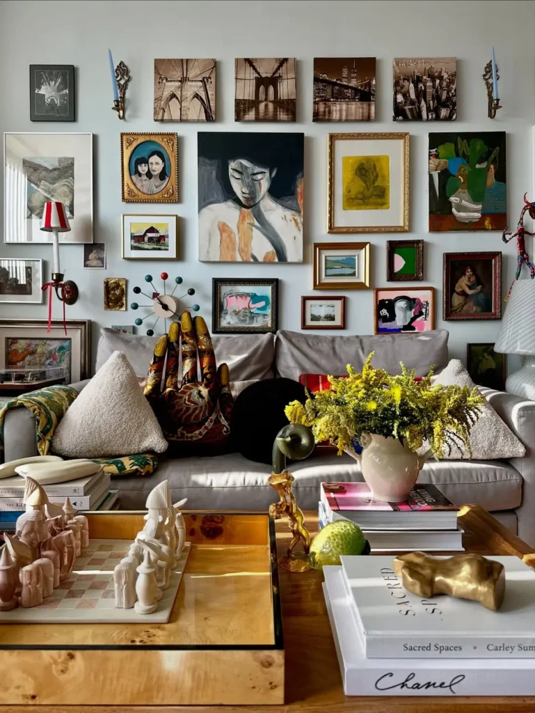 Maximalist eclectic living room with a gray sofa, patterned pillows, art-filled gallery wall, a coffee table topped with books, flowers, a chess set, and various decorative objects.