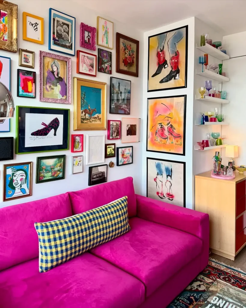 A bright pink couch sits against a wall covered with colorful framed art, adding to the maximalist vibe. Shelves with glassware and a wooden dresser are on the right.
