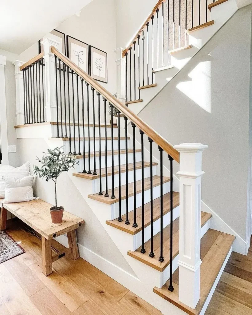 A wooden staircase with black metal balusters and white trim, next to a wooden bench with a potted plant and framed artwork on the wall.