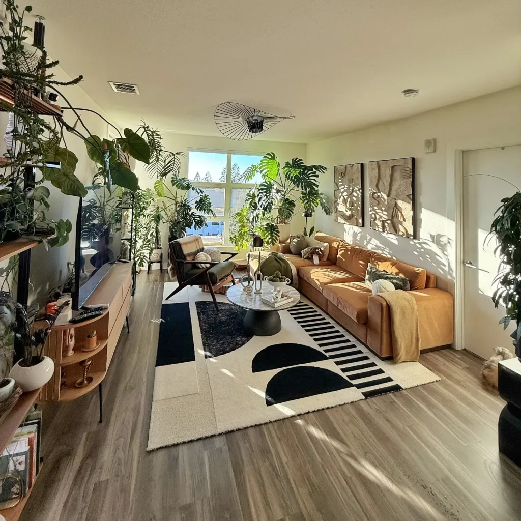 A sunlit living room with a brown sofa, black-and-white rug, modern coffee table, mid century modern chairs, indoor plants, wall art, and wood flooring. Large windows let in natural light.