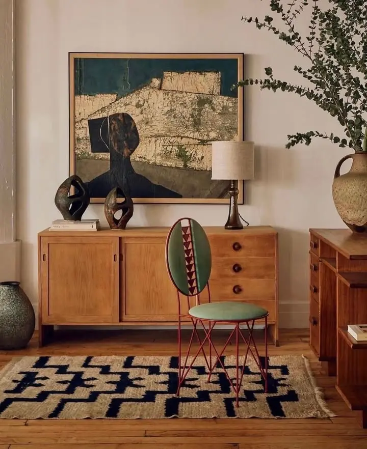 A stylish mid century modern room featuring a wooden sideboard, abstract art, sculptural decor, a green chair, a patterned rug, and large vases with branches.