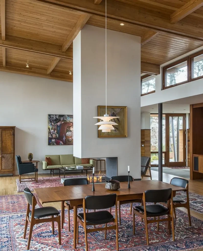 A modern dining room with a wooden table, six chairs, mid century modern pendant light, and large rugs; art and a fireplace divide the open-plan space with visible wood ceiling beams.