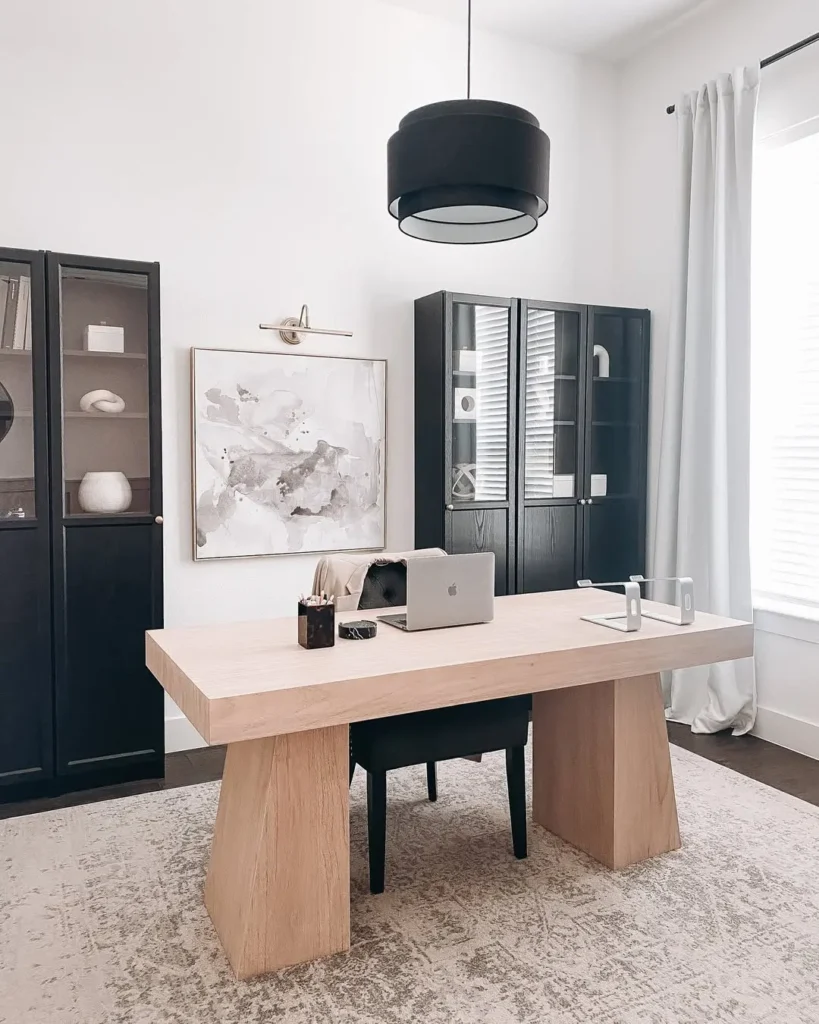 A minimalist home office featuring a light wood desk, black chair, dark display cabinets, abstract wall art, and a laptop on the desk, all illuminated by a striking black pendant light.