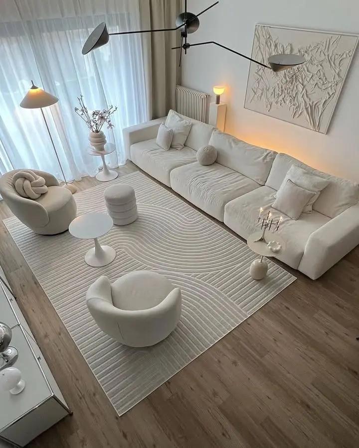 Minimalist modern living room with white furniture, textured white rug, abstract wall art, and wooden floor, lit by natural light from large windows and a stylish black ceiling lamp.