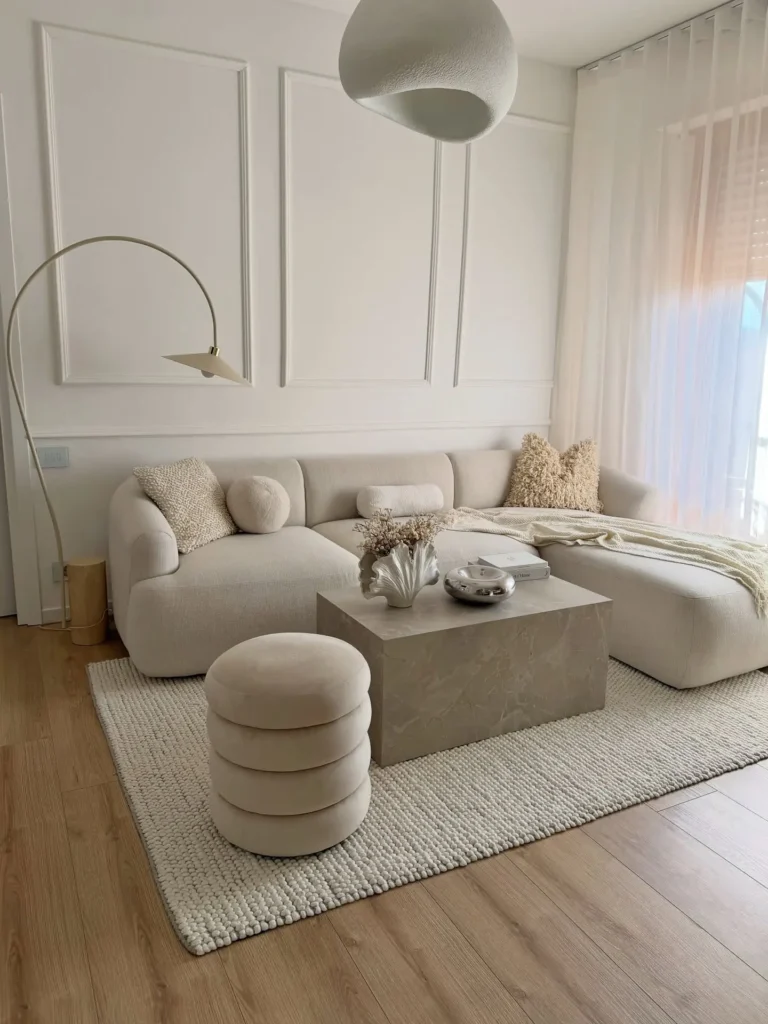 Modern minimalist living room with a white sectional sofa, textured cushions, a round ottoman, marble coffee table, arc floor lamp, and neutral decor arranged on a rug over wood flooring.