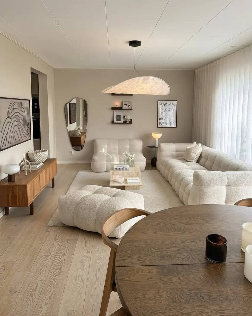 Modern living room with cream-colored sectional sofas, a wooden coffee table, abstract wall art, a round dining table, and large windows with sheer curtains in minimalist style and calming colors.