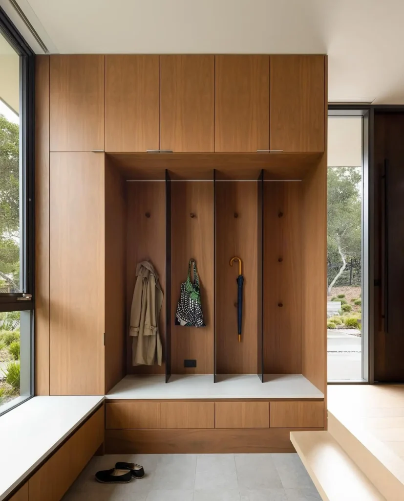 A minimalist modern wooden mudroom with three open cubbies, a coat, umbrella, and bag hanging, drawers below, tall cabinets above, a window seat, and shoes on the floor.