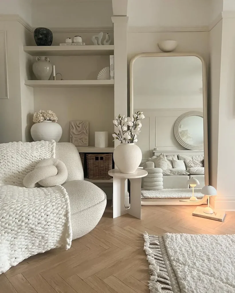 A minimalist neutral living room with a curved white armchair, textured blanket, large mirror, decorative vases, and a rug on a wooden floor.