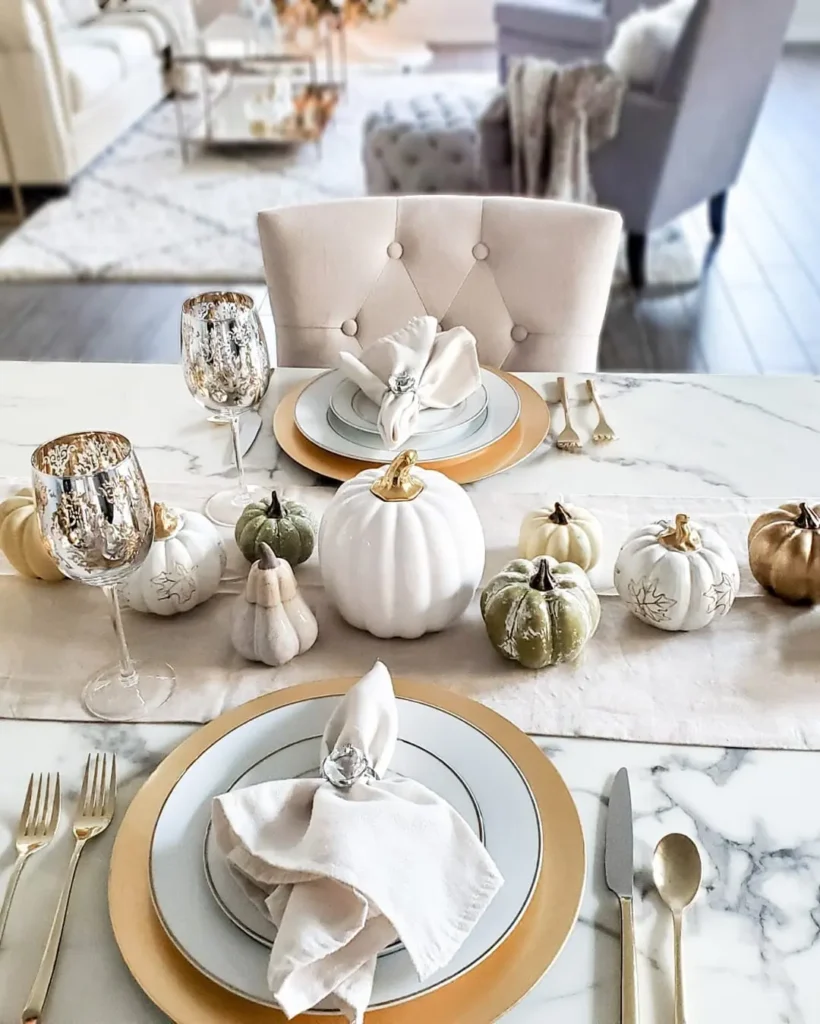 A marble minimalist thanksgiving dining table set with gold-rimmed plates, napkins, gold cutlery, and decorative pumpkins in neutral tones, with plush chairs and a living area in the background.
