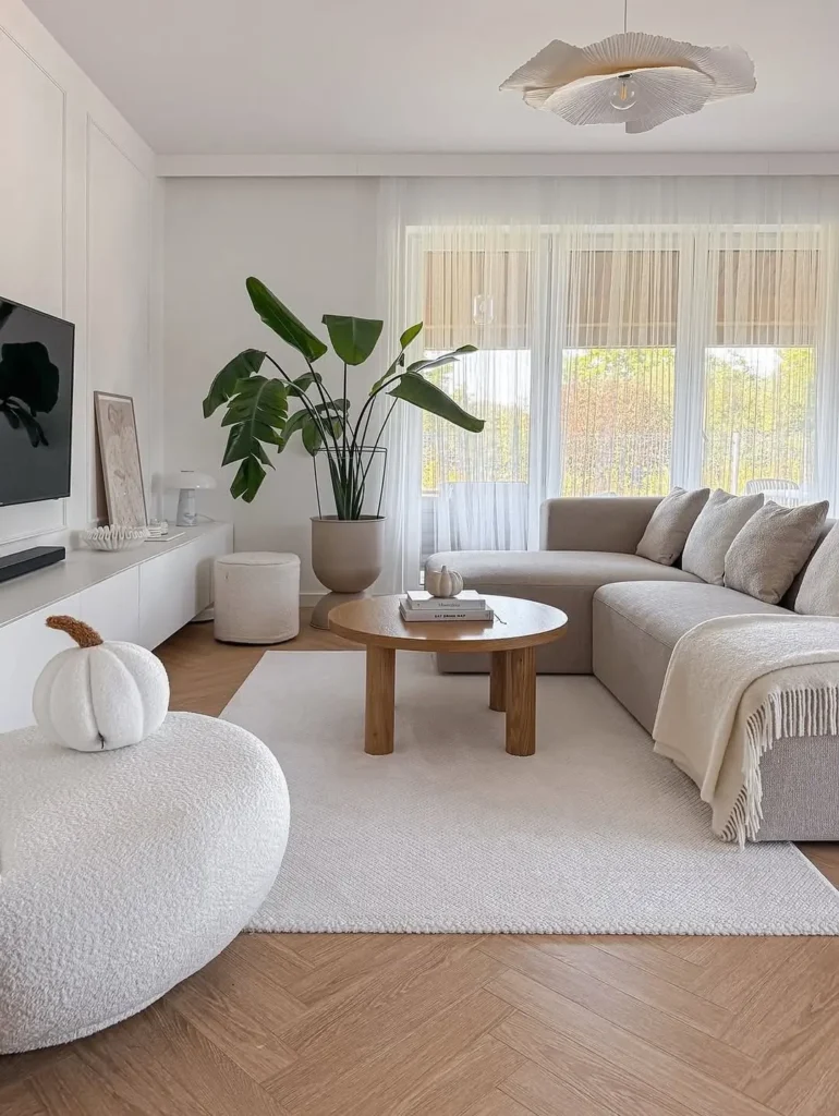 A minimalist living room with a beige sectional sofa, round wooden coffee table, large indoor plant adding a natural element, wall-mounted TV, and light-colored decor on a herringbone wood floor.