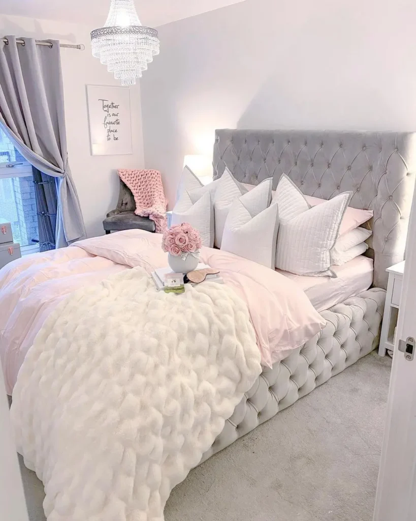 A neatly made bed with tufted headboard, layered pillows, pink bedding, a white textured throw, bedside tables, a chair, and a chandelier in a bright, modern bedroom.