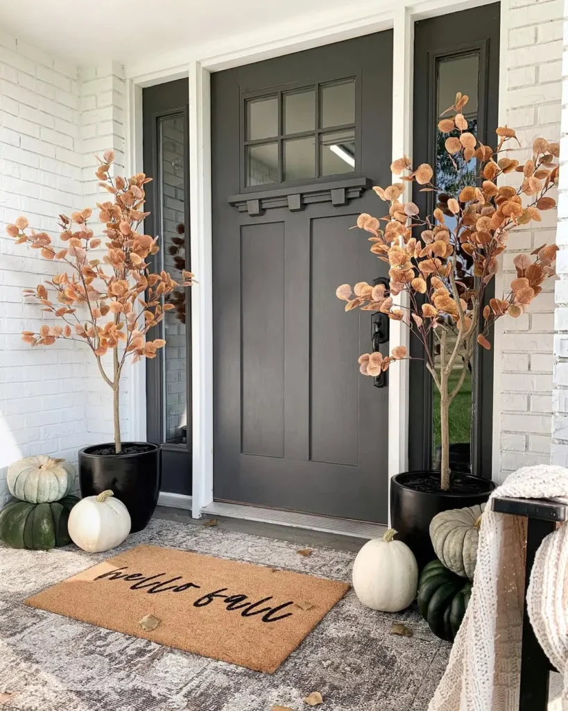 A modern fall front porch featuring a dark gray door, potted autumnal plants, white and green pumpkins, and a “hello fall” doormat layered on a patterned rug.